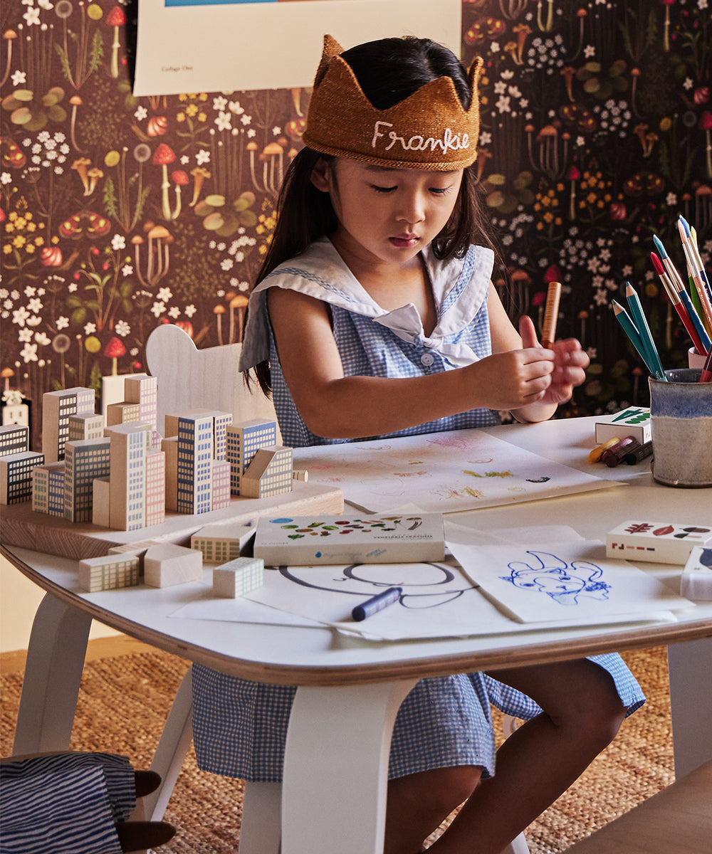 A child wears a crown, drawing at a desk with art supplies and Le Manhattan Skyline Blocks on a patterned background.