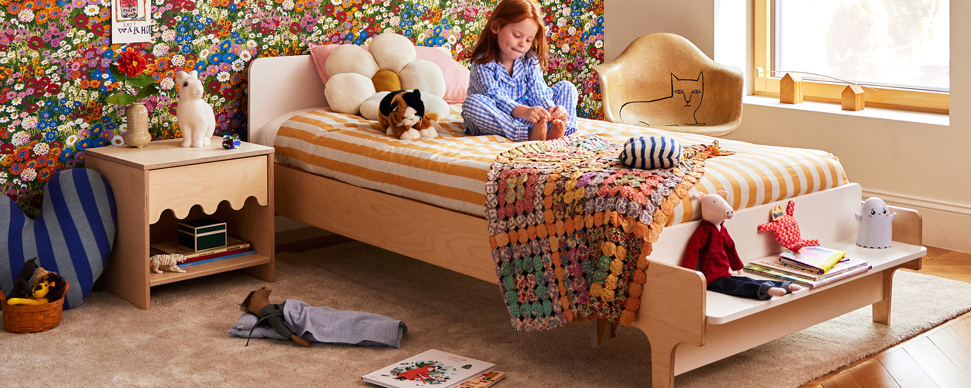 Child sitting on a bed in a colorful, cozy bedroom with a floral wall, toys, and books scattered around. A small nightstand and a cat-shaped chair are nearby.