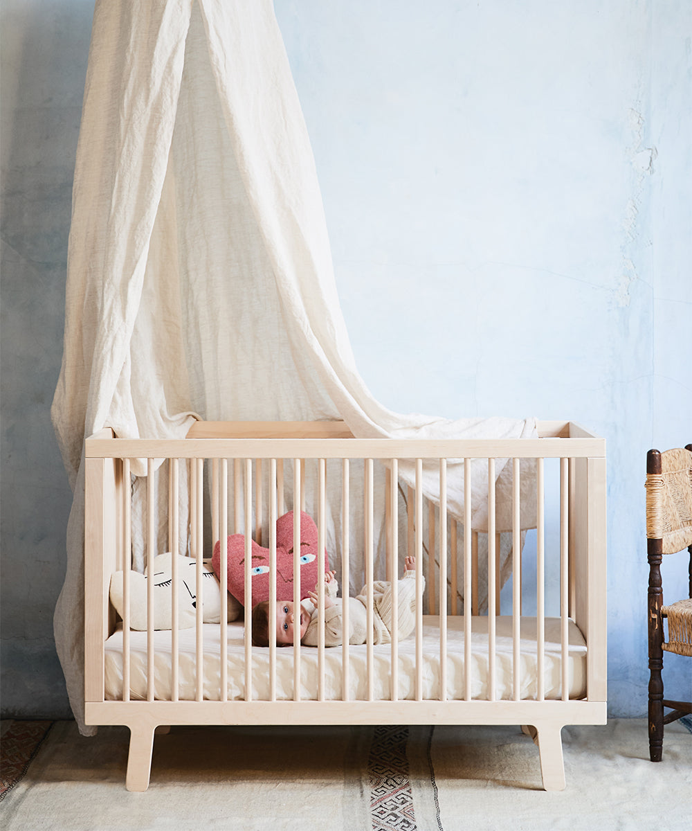 A wooden crib with a beige canopy holds a baby and two stuffed toys on the eco-friendly Dual Firm Crib Mattress. A soft blue wall forms the background, while a wooden chair is partially visible on the right.