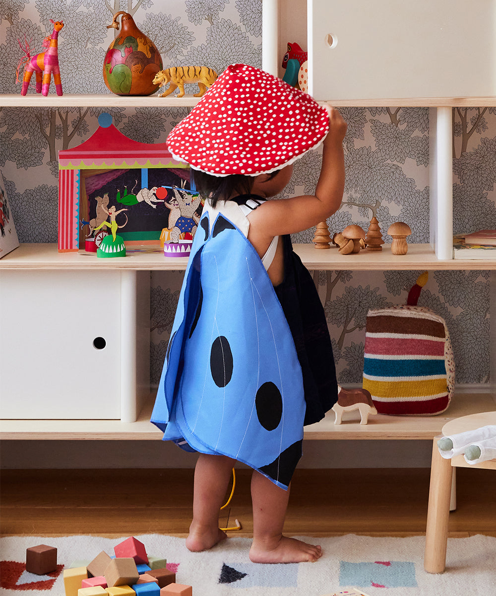 A child in Le Blue Butterfly Wings and a red mushroom hat plays in a colorful room filled with toys and books.