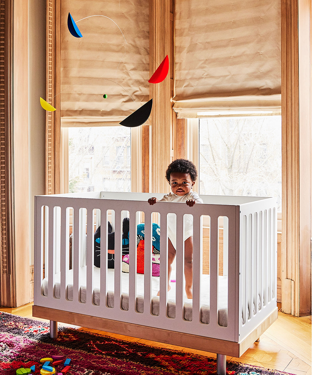 A baby stands in a white crib in a room with large windows, playful toys scattered on the floor, and the colorful Le Mobile - Life & Thread hanging above.