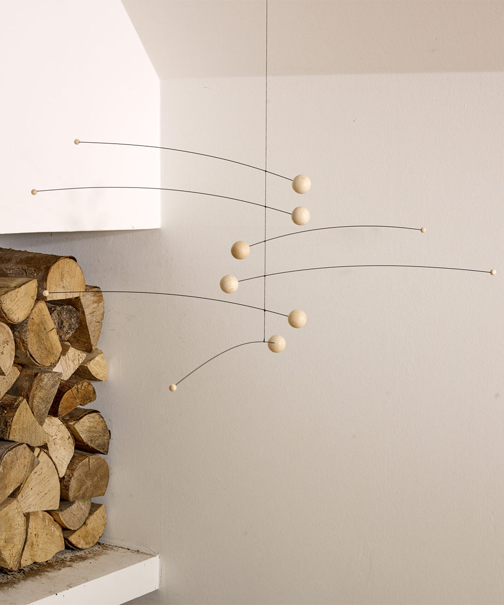 The Le Mobile - Futura Nature, a minimalist mobile with wooden spheres, hangs near stacked firewood against a white wall.