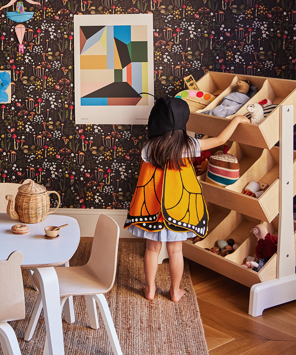 A child wearing a Le Antennae Hat reaches for toys on a shelf in a colorful, decorated room with a small table and chairs.