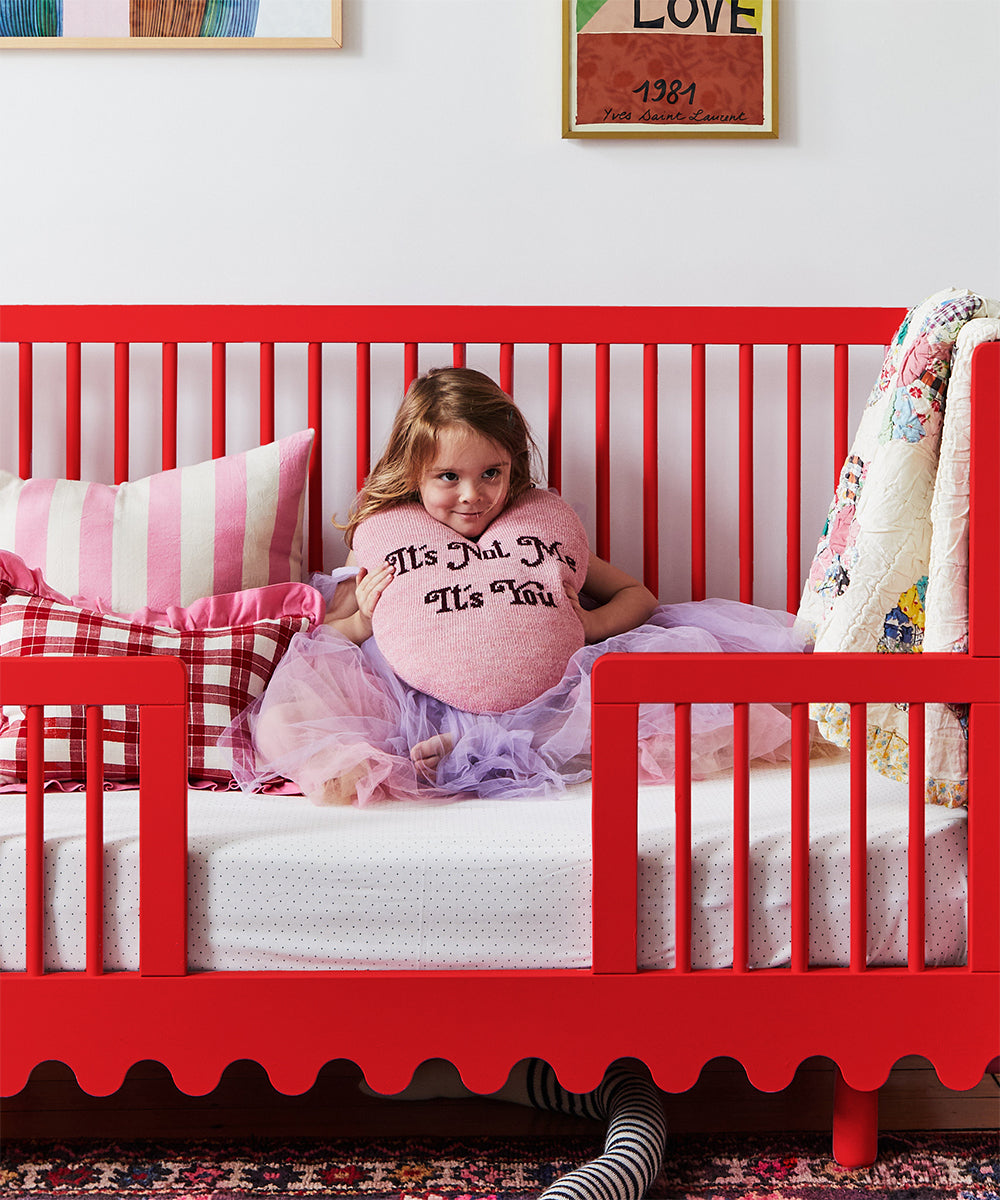 A child sits in a crib upgraded with the Moss Crib Conversion Kit, holding a heart pillow while wearing a tutu. The scene is enhanced by vibrant bedding and decorative wall art in the background.