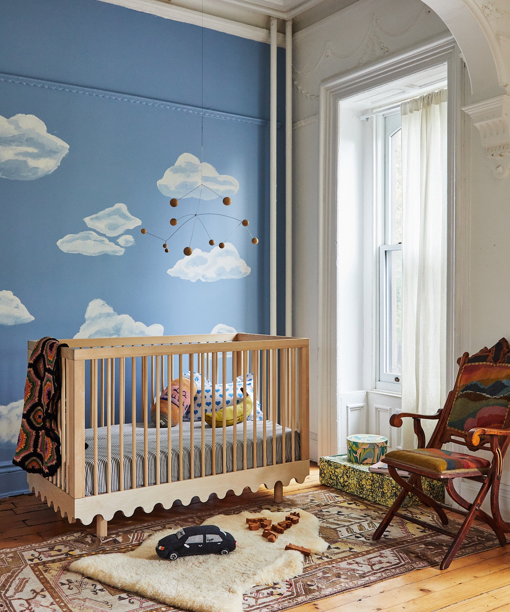 Baby room with a cloud mural, wooden crib, My Ride Pillow, and wooden toys on a shag rug near a window.