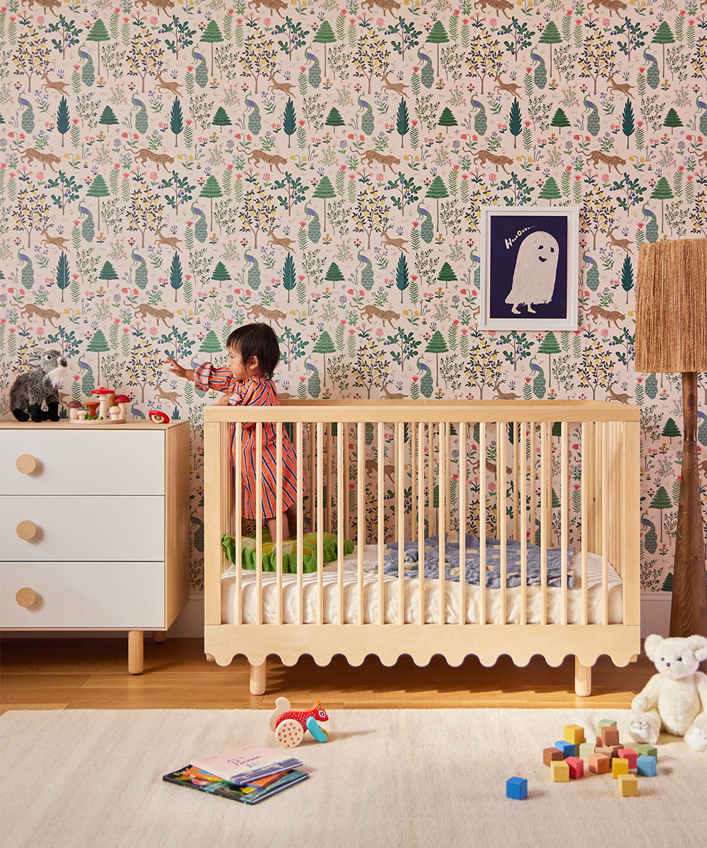 A child in a Moss Crib reaching for toys on the dresser, surrounded by colorful nature motif wallpaper and soft toys scattered on the floor.