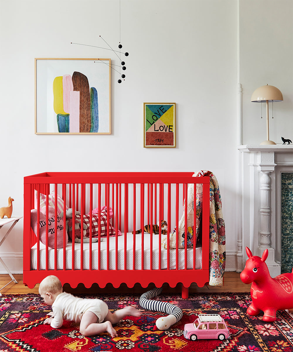 A baby is crawling near a Moss Crib, surrounded by colorful toys and art in a nursery with a vibrant carpet.