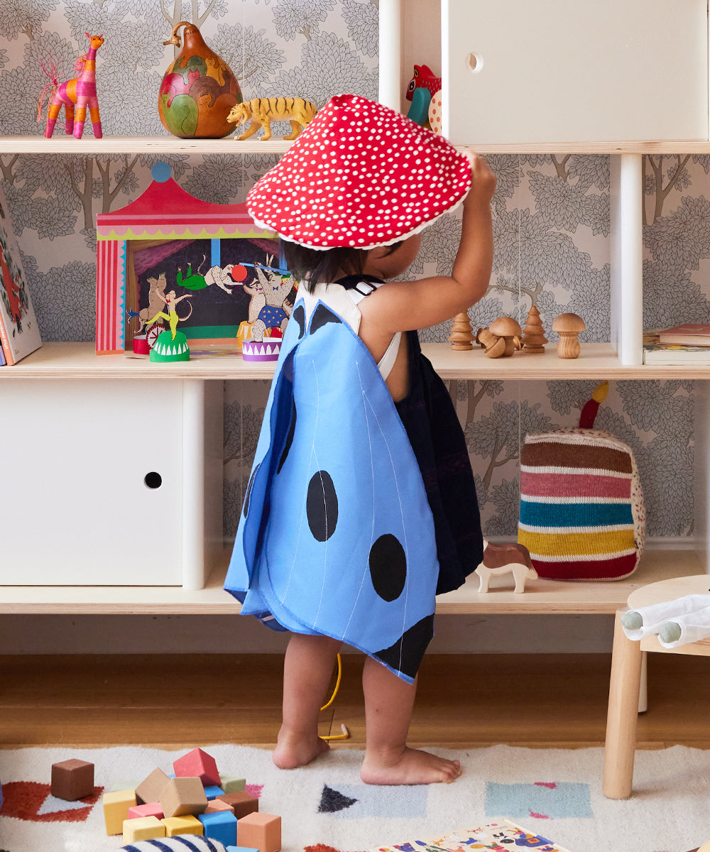 A child wearing Le Mushroom Hat in red polka-dots and blue ladybug wings stands in a playroom with toys and a shelf in the background.