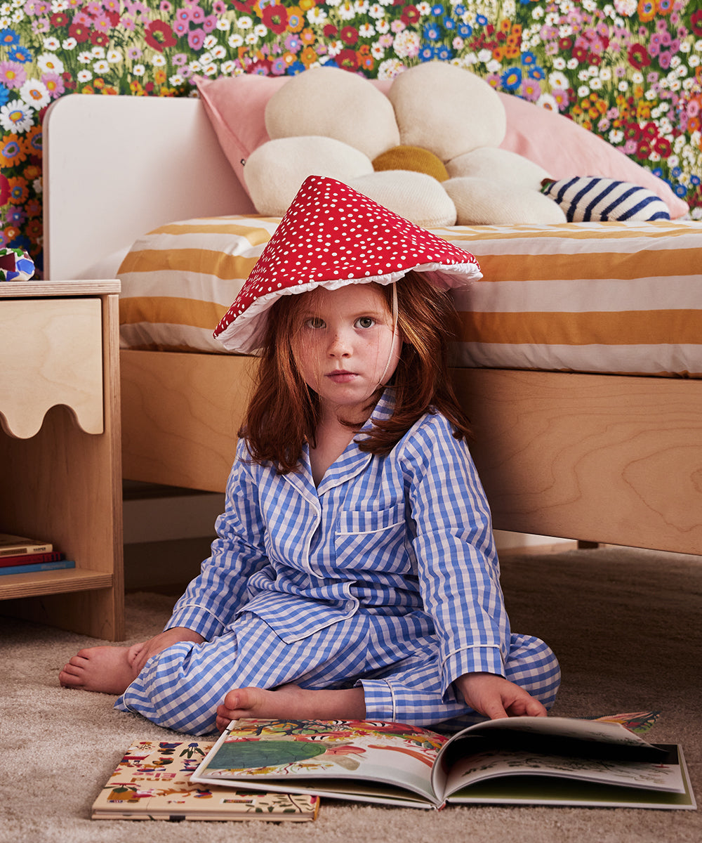 A child in blue pajamas sits on the floor with an open book, wearing the Le Mushroom Hat. The bedroom has floral wallpaper and a bed with striped bedding and a flower-shaped pillow.