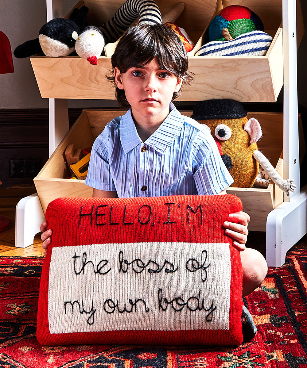 Child holding a Name Tag Pillow with "HELLO, I'M the boss of my own body" sits among stuffed toys.