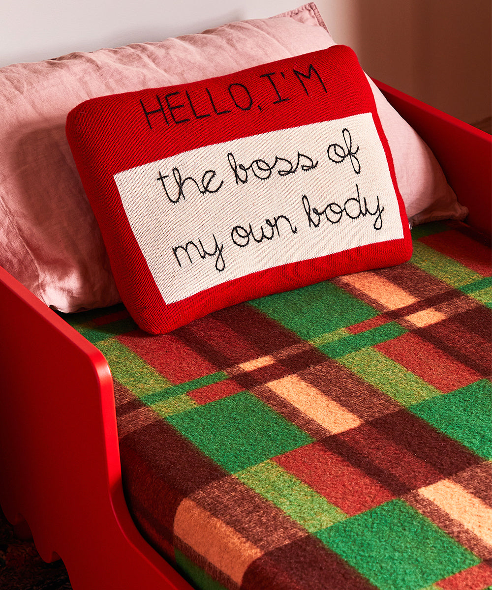 Red bed featuring a colorful plaid blanket and a Name Tag Pillow with "HELLO, I'M the boss of my own body" in black script.