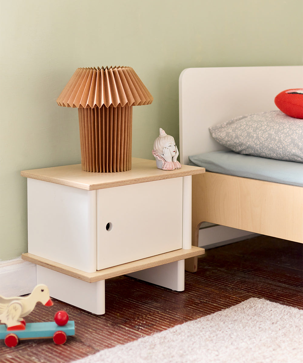 ML Nightstand with a paper lamp and figurine beside a bed; toy duck on wheels on the floor.