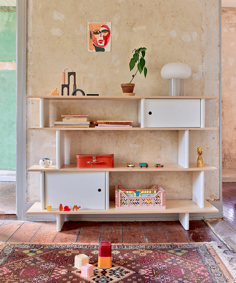 The Mini Library holds books, toys, and decor against a textured wall, with a rug and toys on the wooden floor.