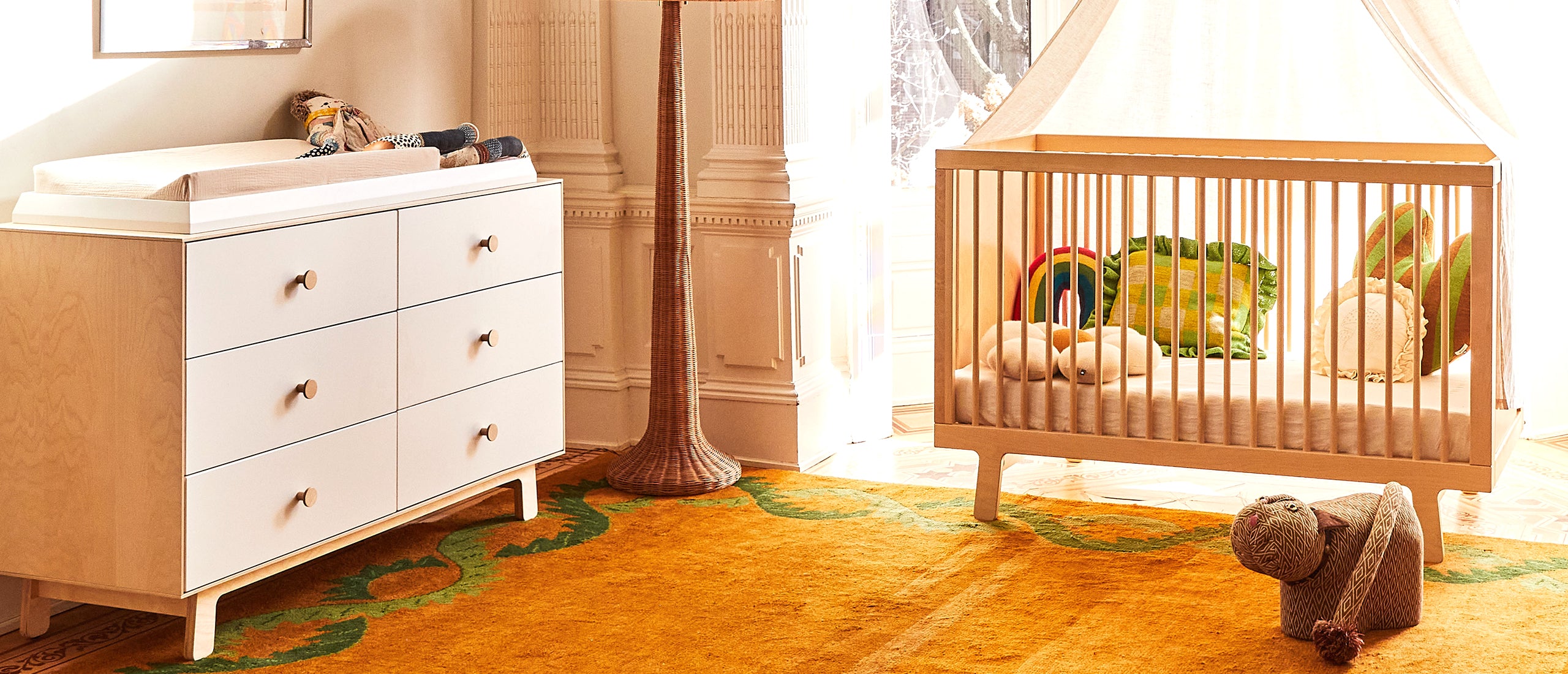 A nursery with a wooden crib, white dresser, patterned carpet, and plush toys on the floor and crib.