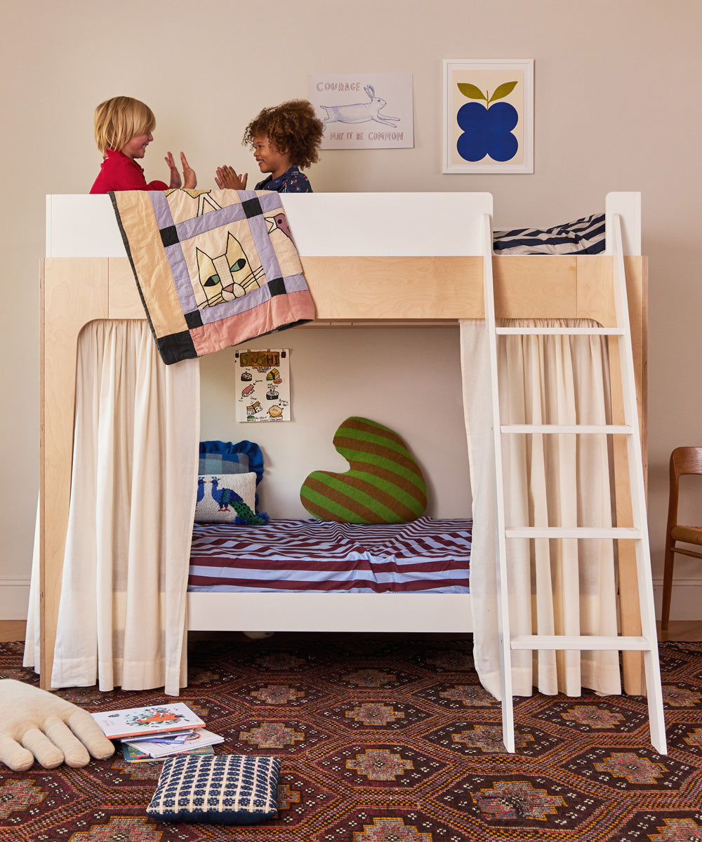 Two children play hand-clapping games on the top bunk of a wooden bunk bed, decorated with a quilt and curtain. The lower bunk holds books and a pillow, while the wall features vibrant Le Indigo Fruit Posters.