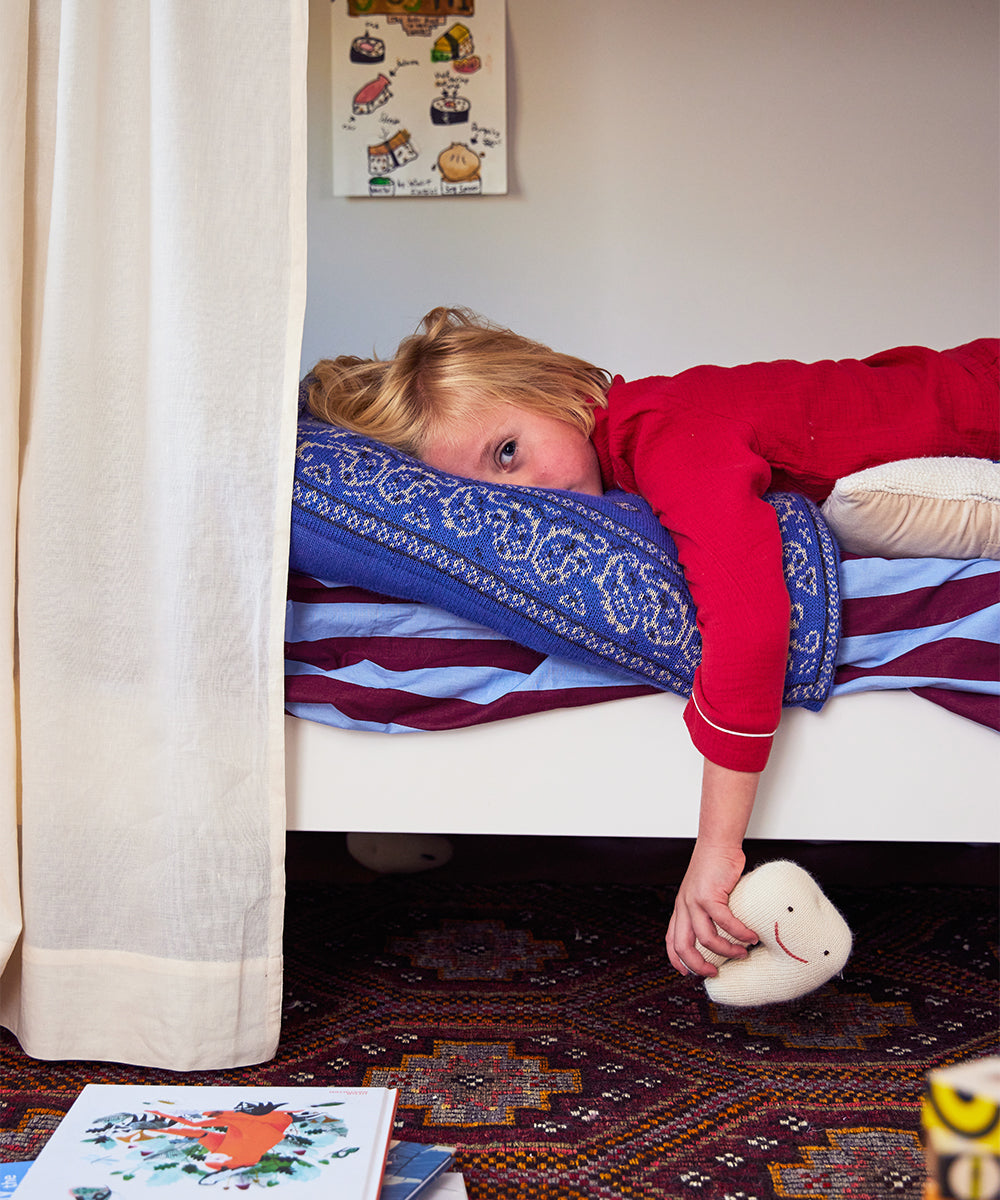 Child in red pajamas holds a Tooth Fairy Pillow on the bed, with books scattered on the floor nearby.