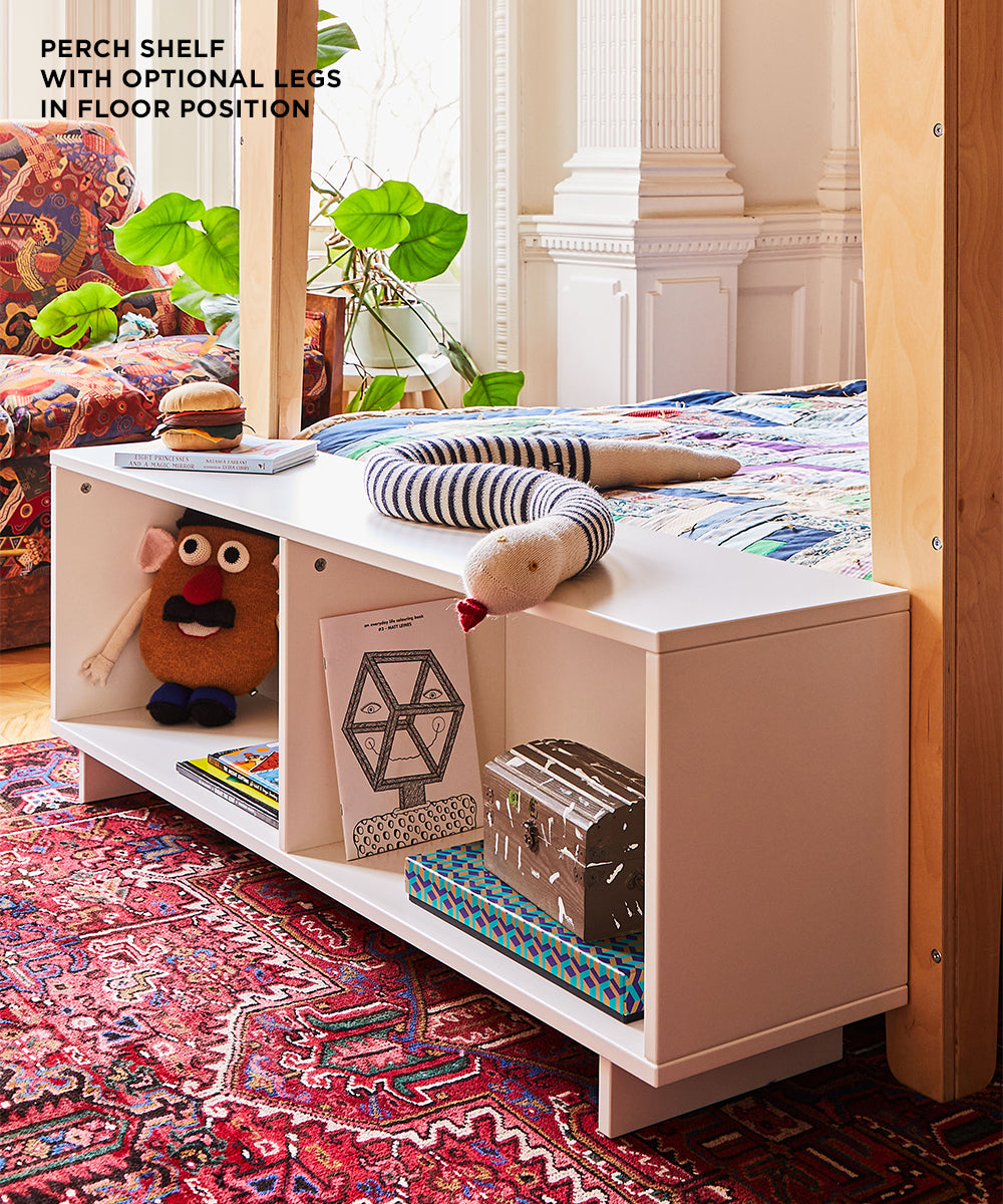 Childrens bedroom featuring the Perch Shelf - Full Size in white at the beds foot, adorned with toys and books. A colorful rug enhances the decor, complemented by a nearby large plant.