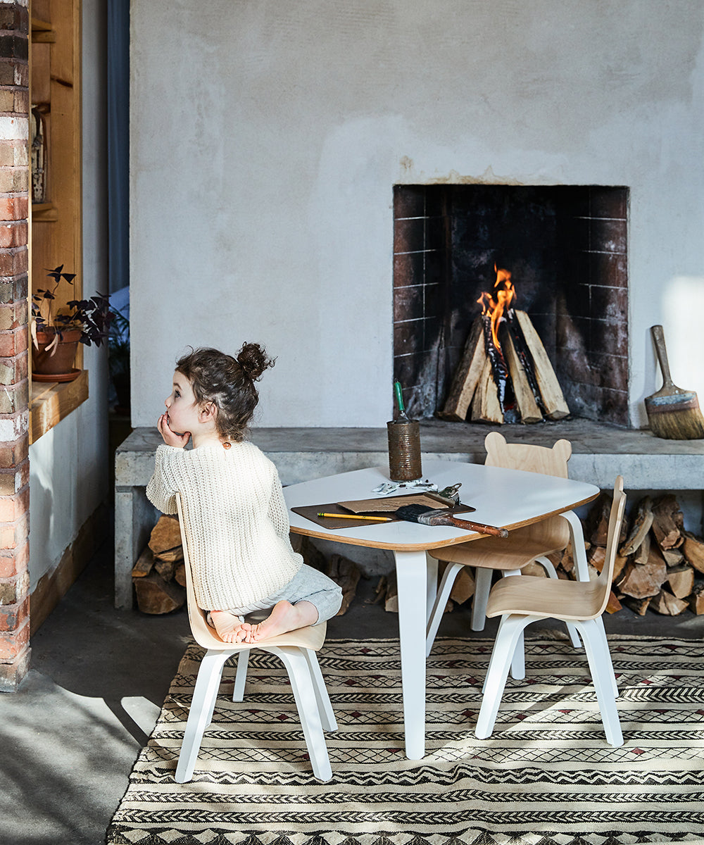 A child sits on one of the Bear Play Chairs from the set of 2, by a small table, gazing away in a room with a lit fireplace and stacked firewood.