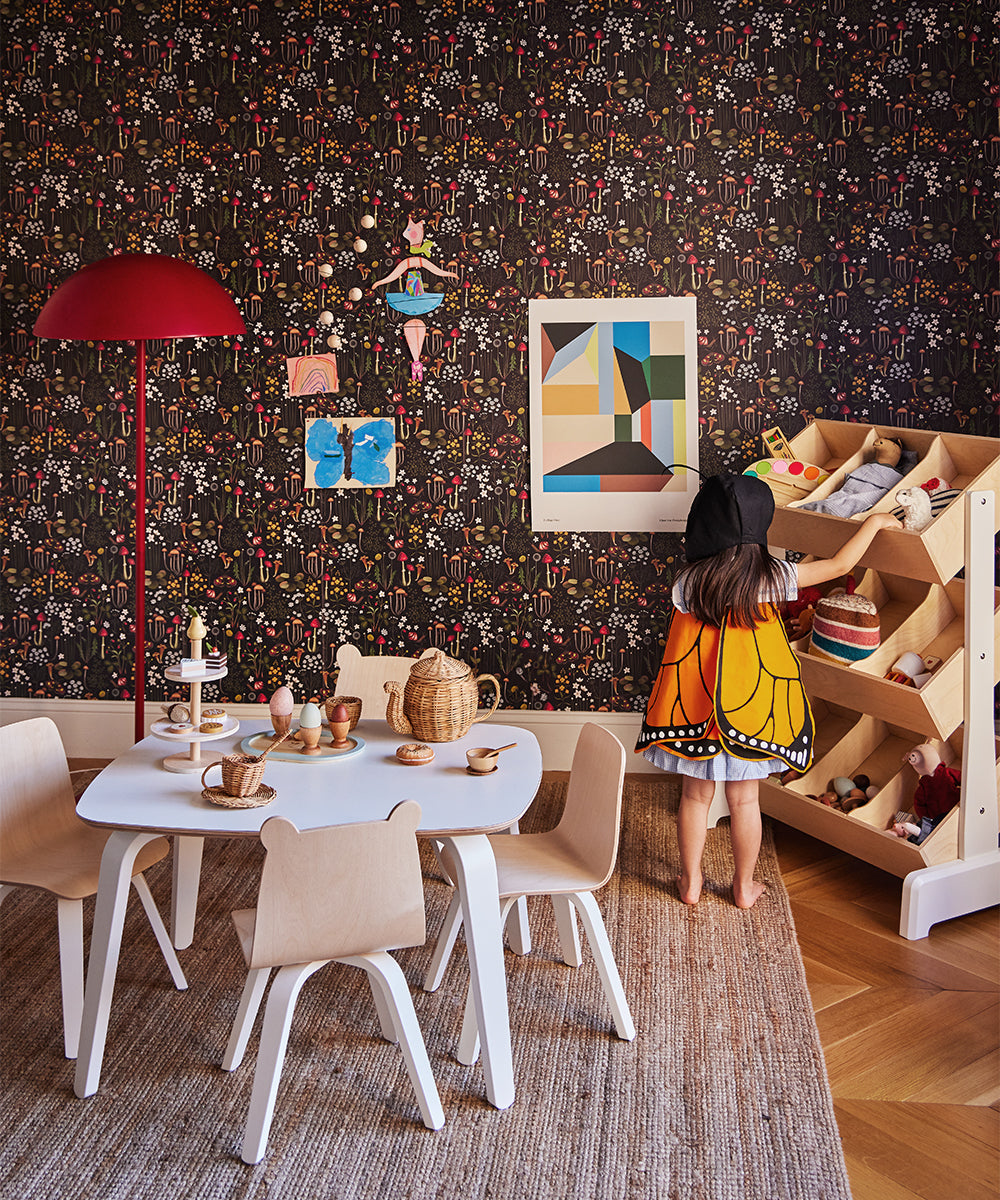 Child dressed as butterfly in playroom with table, chairs, Le Wooden Dish Set, toys, and patterned wallpaper featuring decorative art.