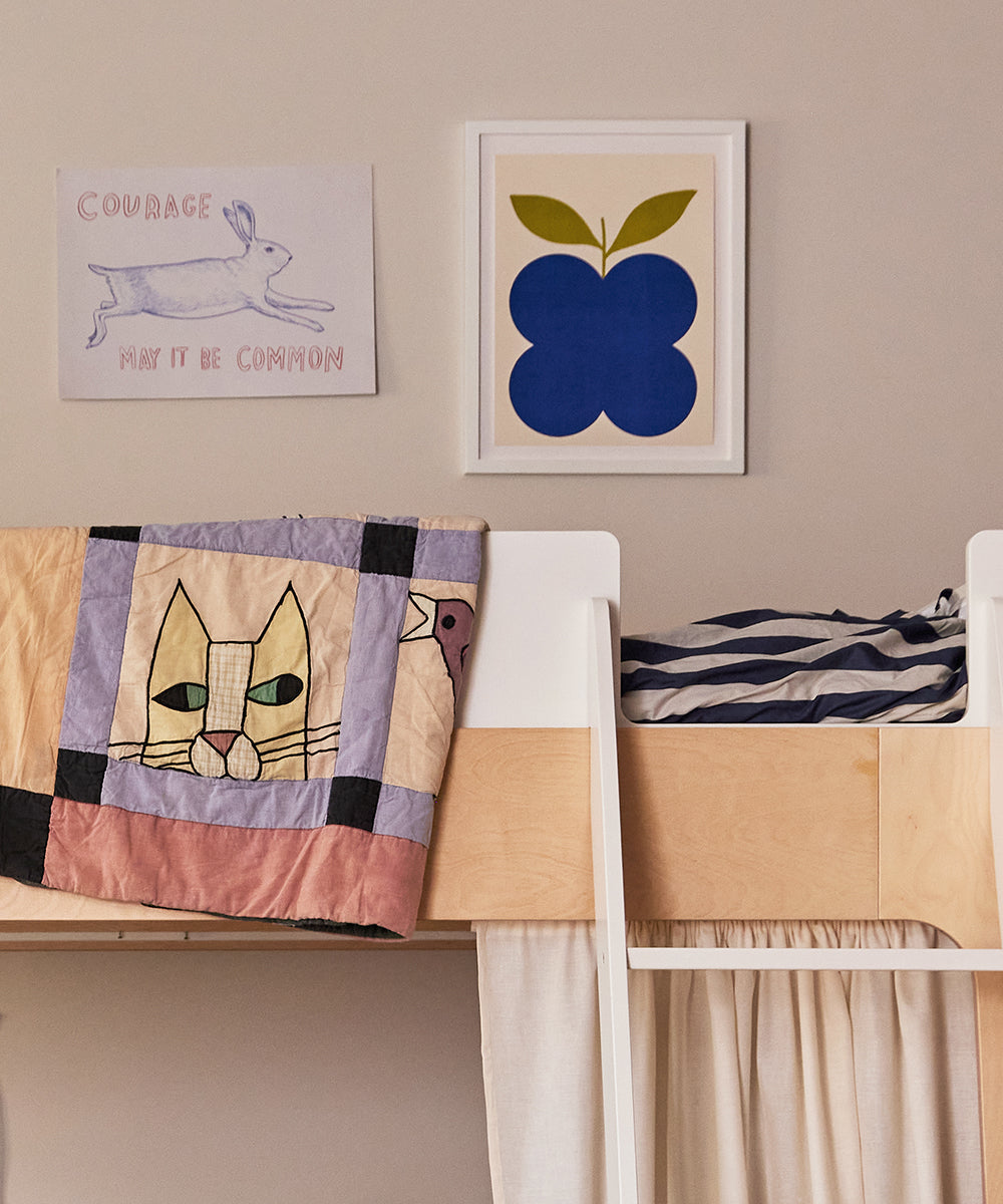 A childs bedroom features a wooden bunk bed with colorful cat-themed quilts. Striped bedding is visible, and two framed art pieces adorn the wall: a rabbit sketch and Le Indigo Fruit Poster showcasing a blue abstract shape.