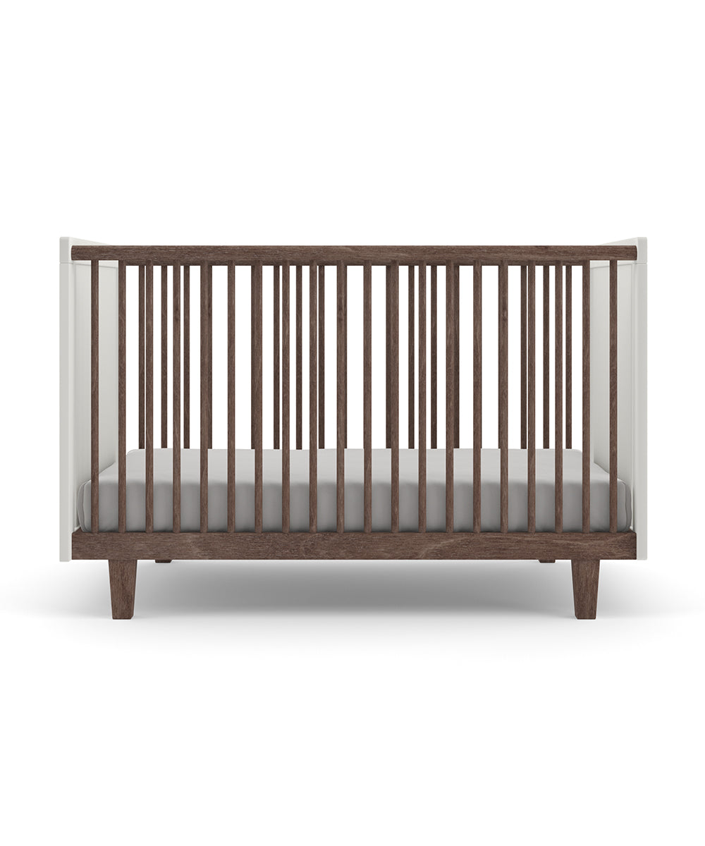 The Rhea Crib is a modern wooden baby crib featuring slatted sides, a white frame, and a gray mattress.