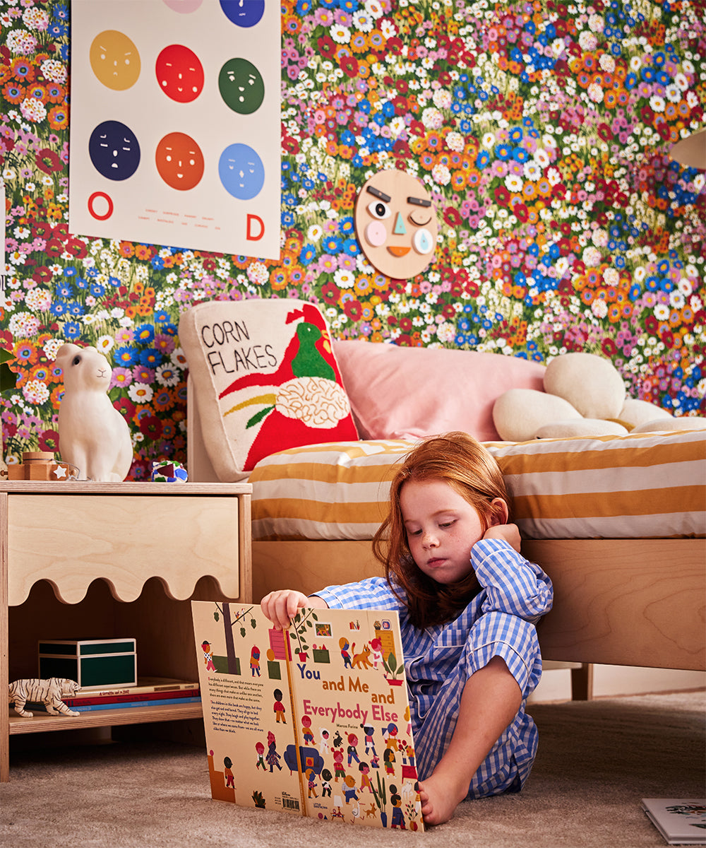 A child in pajamas reads a book in a vibrant floral-themed room with playful decor and relaxes on a plush bed featuring the Corn Flakes Pillow.