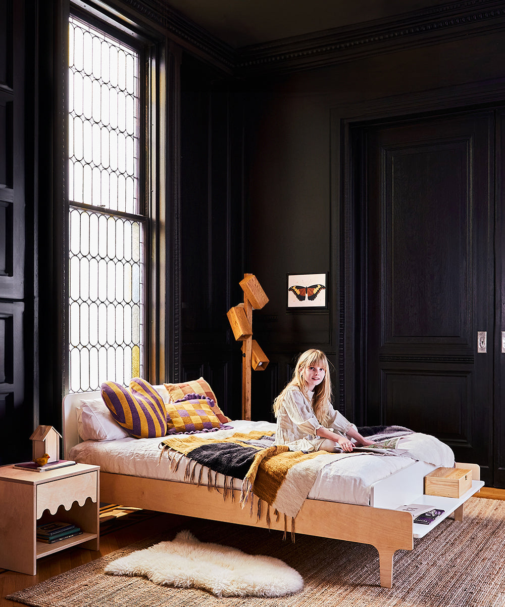 A woman sits on a River Full Bed in a dark-walled room featuring a large window, wooden lamp, and rug.