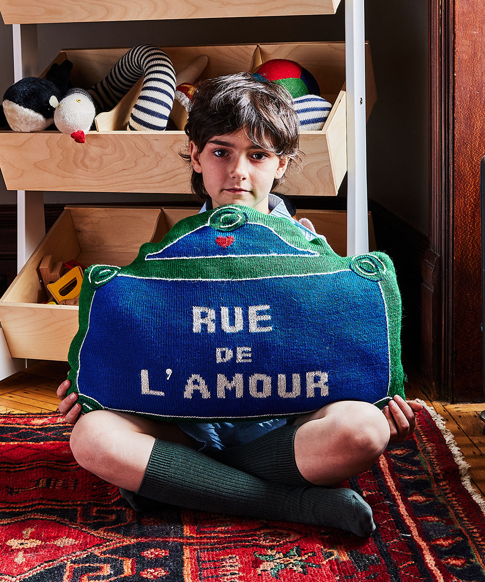 A child sits on a patterned rug holding the Rue de lAmour Pillow, with toys visible in the background.