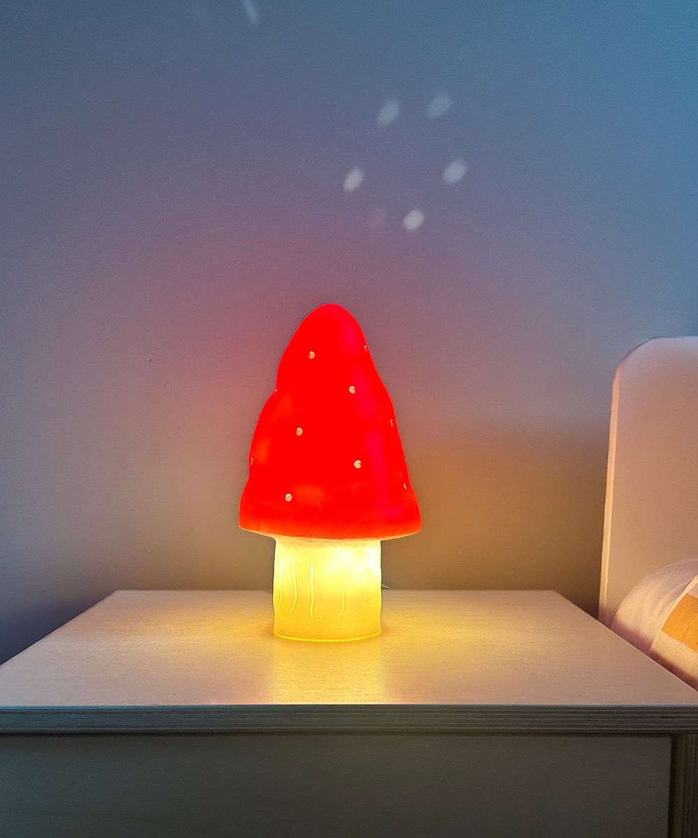 The Le Small Mushroom Lamp, featuring a red cap and white stem, glows on a bedside table against a blue wall.