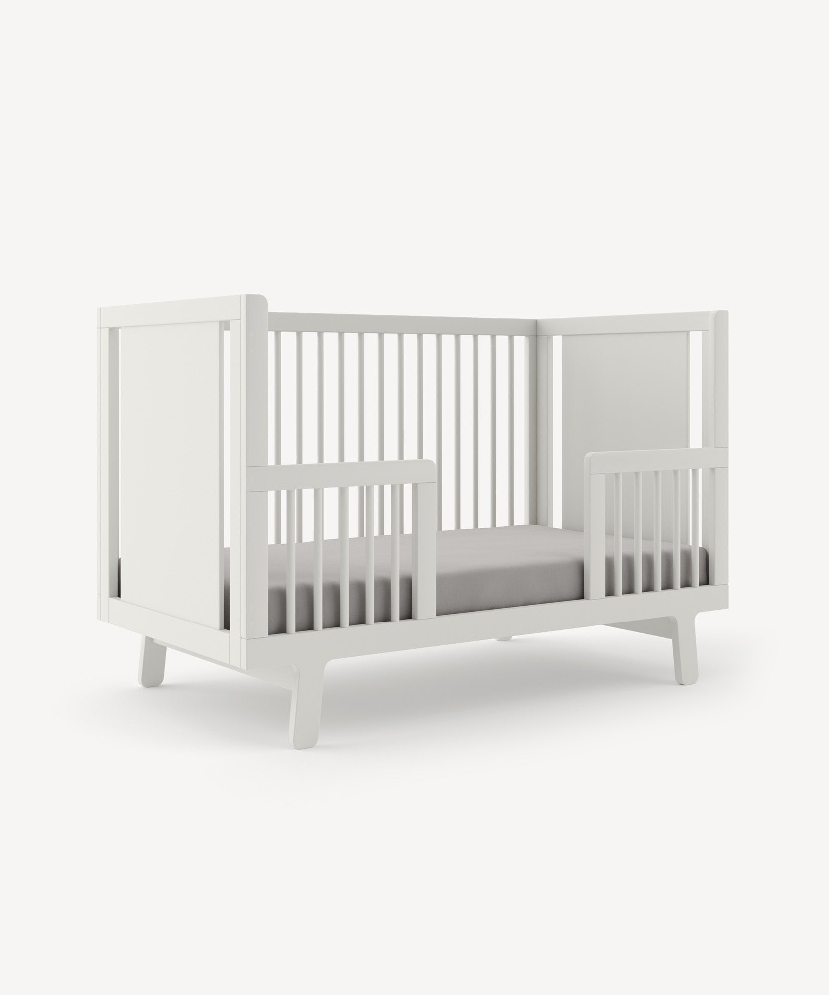 The Sparrow Toddler Bed Conversion Kit is a minimalistic white crib with open slats, a soft mattress, and a plain backdrop.