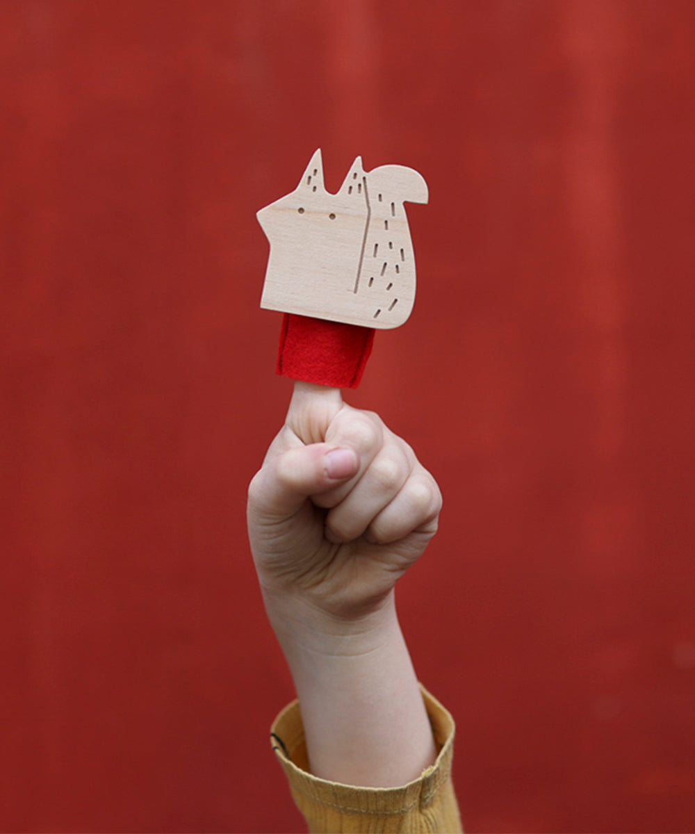 A hand displays a small Le Squirrel Finger Puppet against a vibrant red backdrop.