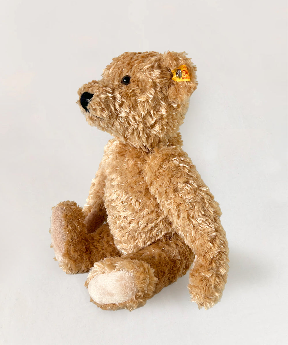 The Le Teddy Bear, Elmar, featuring a yellow tag in its ear, sits upright on a plain white background.