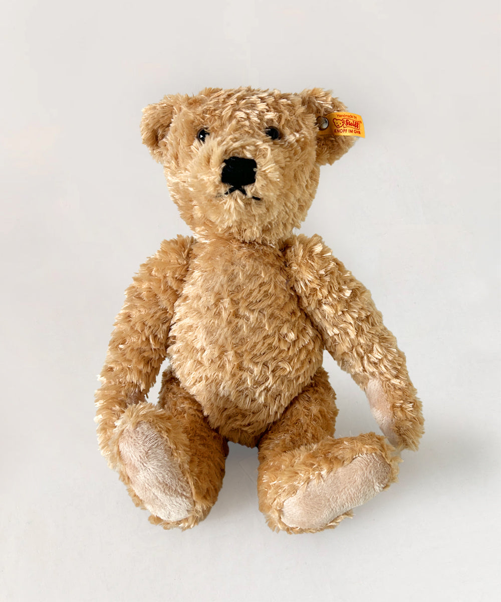 The Le Teddy Bear, Elmar is a fluffy brown bear with a black nose and a brand tag on its ear, sitting upright against a plain background.