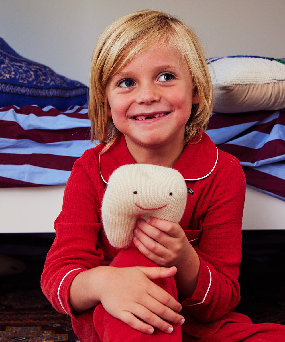 In a bedroom, a smiling child in red pajamas holds a Tooth Fairy Pillow.