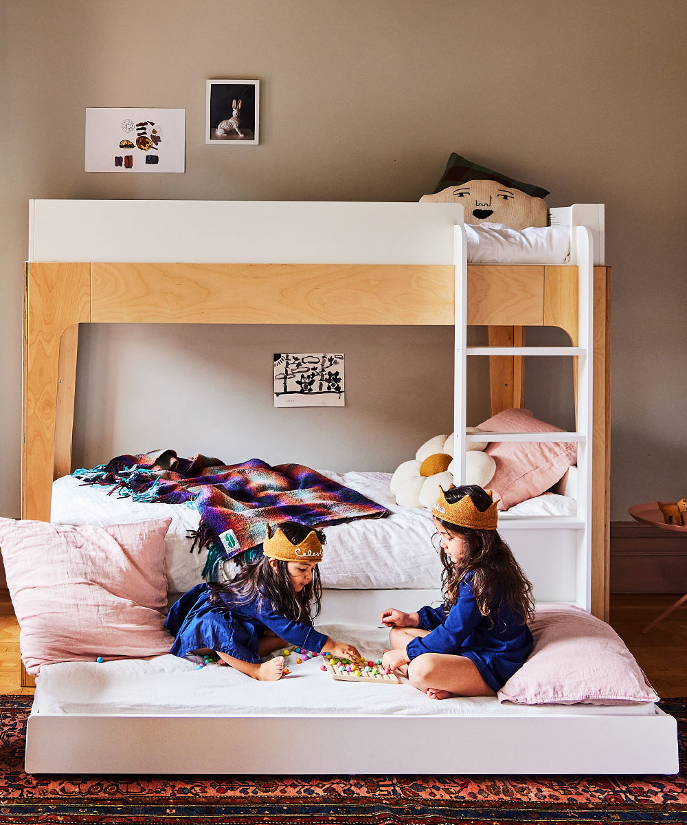 Two children wearing crowns play on a Trundle Mattress under a wooden bunk bed in a cozy room.