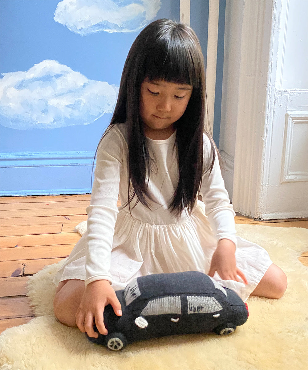 A young girl in a white dress plays with a My Ride Pillow, shaped like a black toy car, on a wooden floor with painted clouds on the wall.