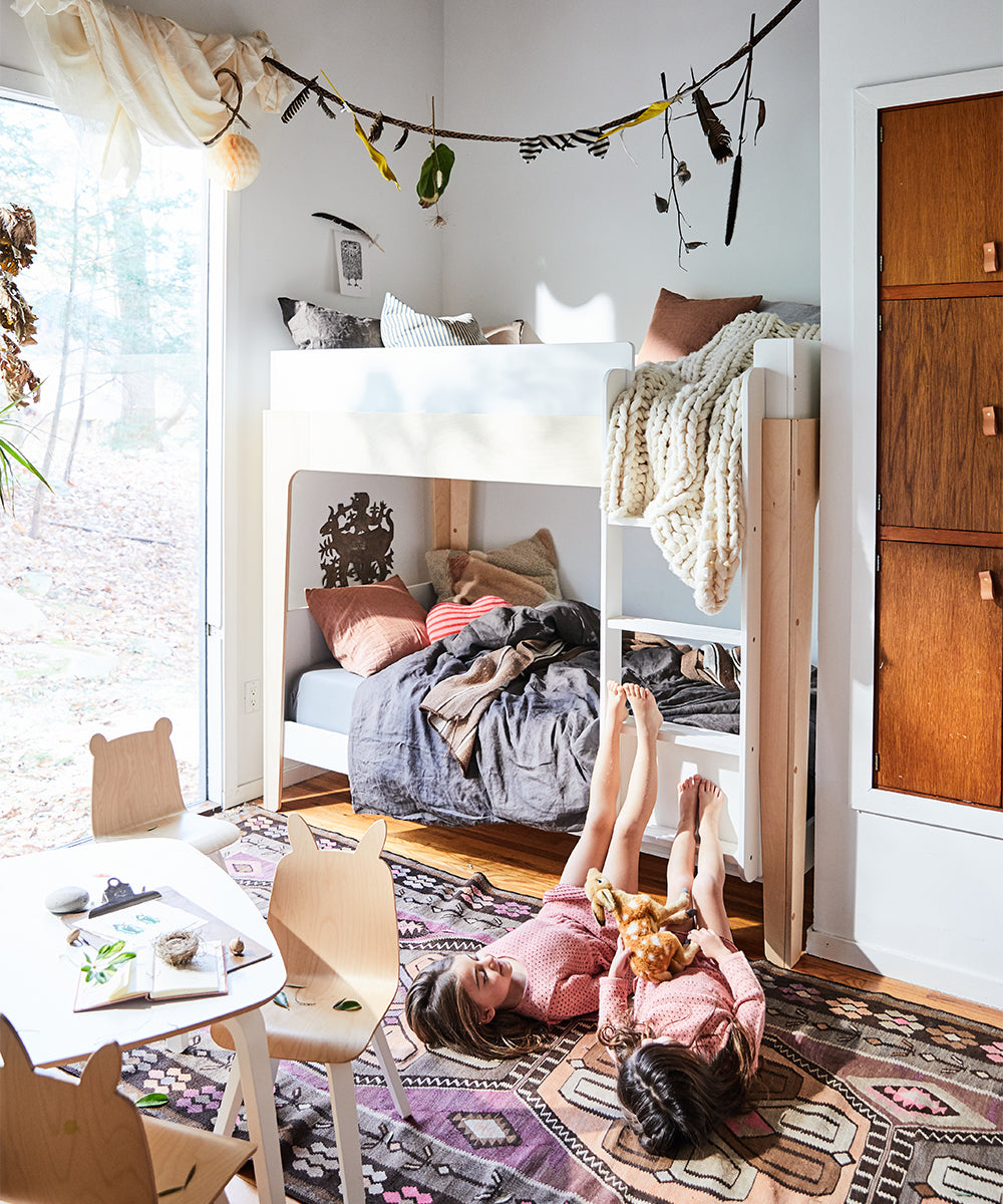 Two children relax on a rug beneath a cozy bunk bed in a room featuring a Perch Ladder Conversion Kit, wooden chairs, a play table, and hanging decor.