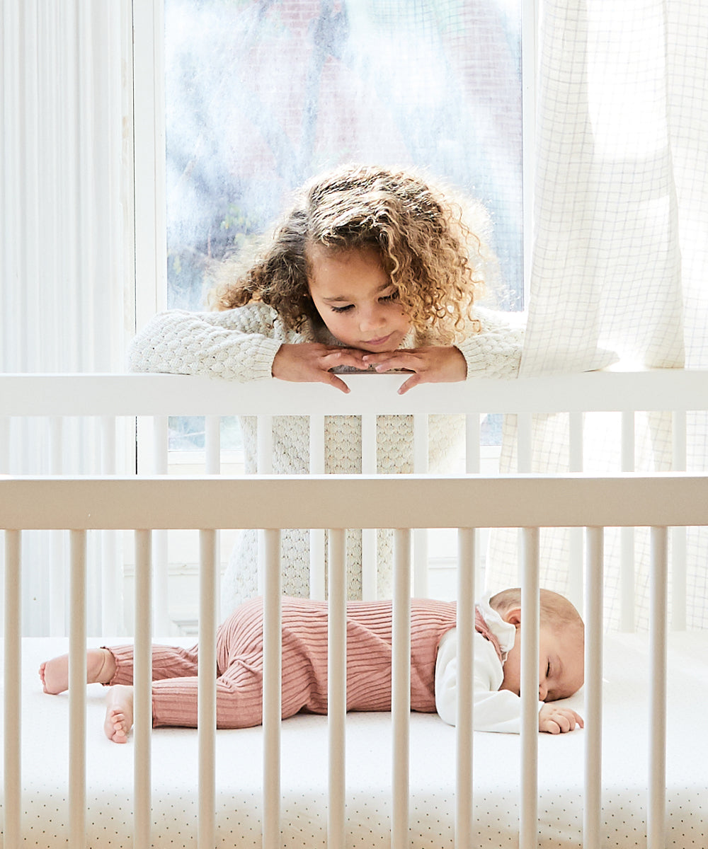 A young girl watches a sleeping baby in a crib, with sunlight pouring through nearby window and curtains. Enjoy peace of mind with our White Glove Service, available exclusively in NYC.