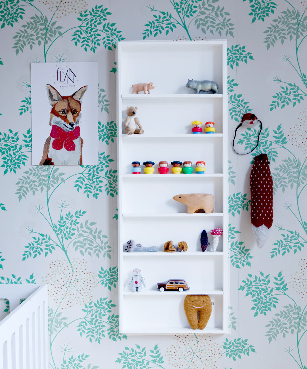 The Convertible XL Changing Station, featuring nursery essentials like toys and a fox illustration, sits next to a plush toy hanging on the wall. A green and brown leaf-patterned wallpaper beautifully enhances this ethically crafted space.