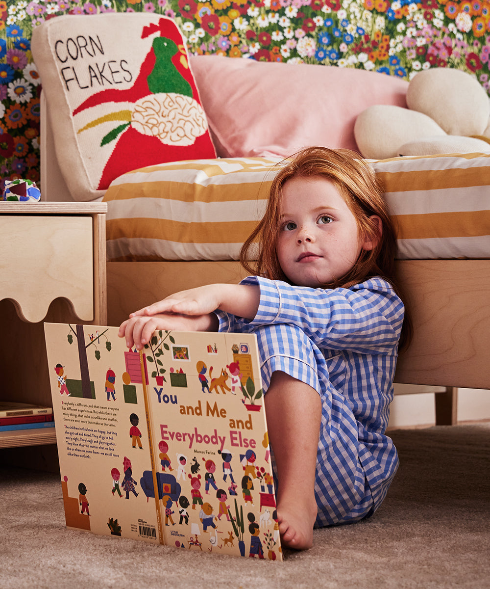 A child wearing blue pajamas sits on the floor by a bed, holding Le Book - You and Me and Everybody Else. A decorative pillow featuring the text Corn Flakes rests on the bed.