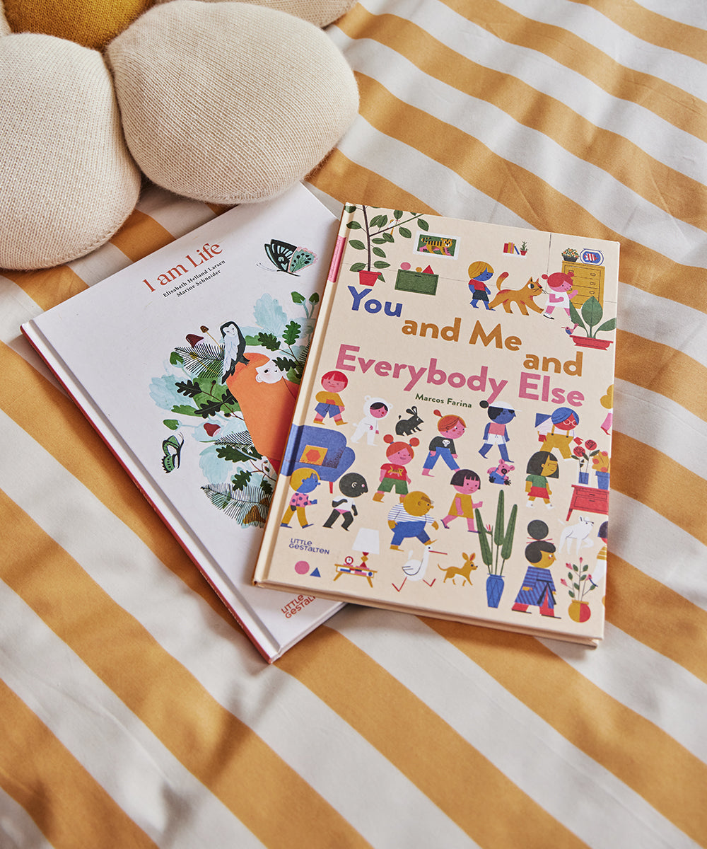 Two childrens books on a striped bedspread: I am Life with an illustrated tree cover, and Le Book - You and Me and Everybody Else featuring colorful illustrations of people and animals.