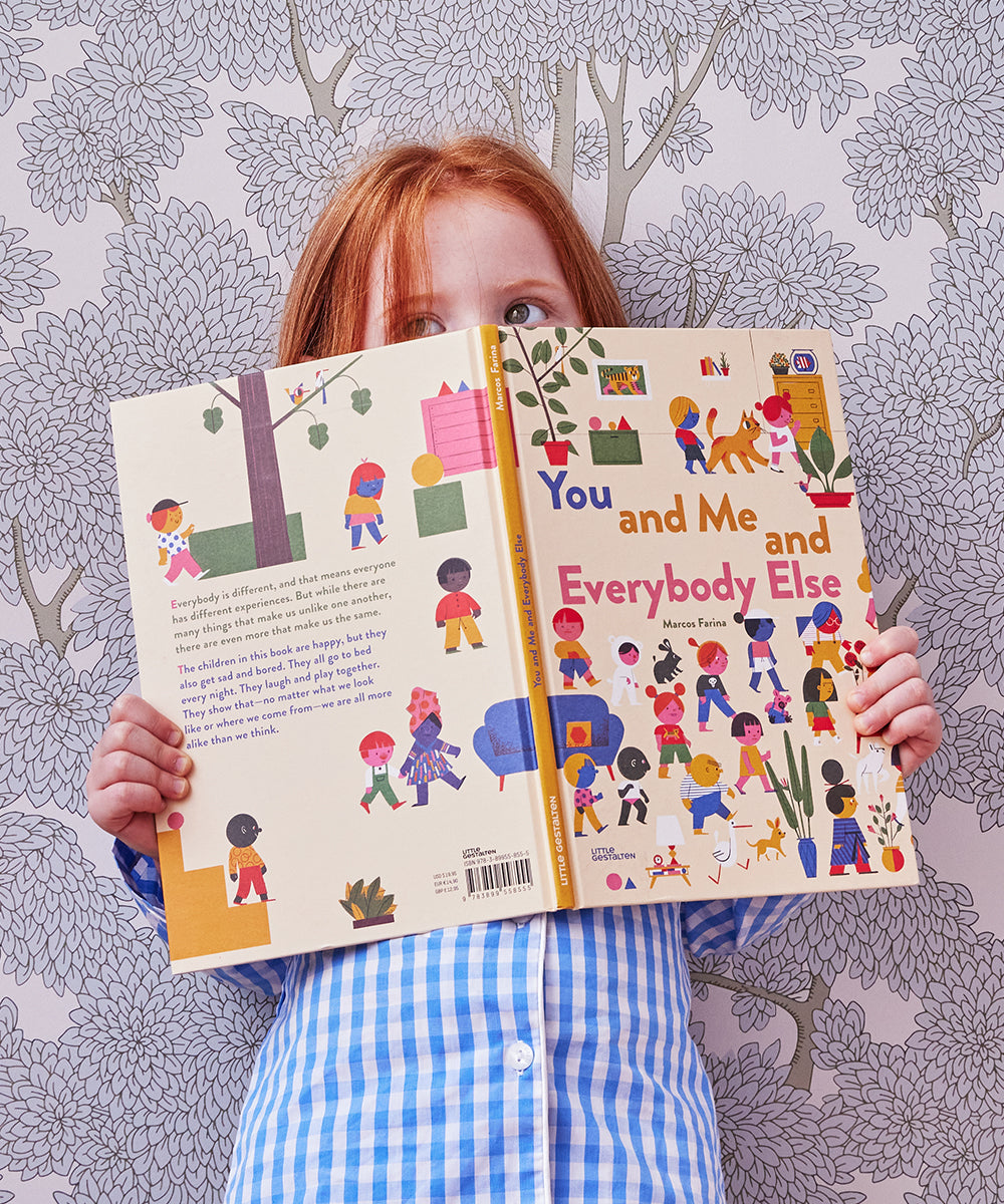 A child holds an open Le Book titled You and Me and Everybody Else in front of a patterned wallpaper background.