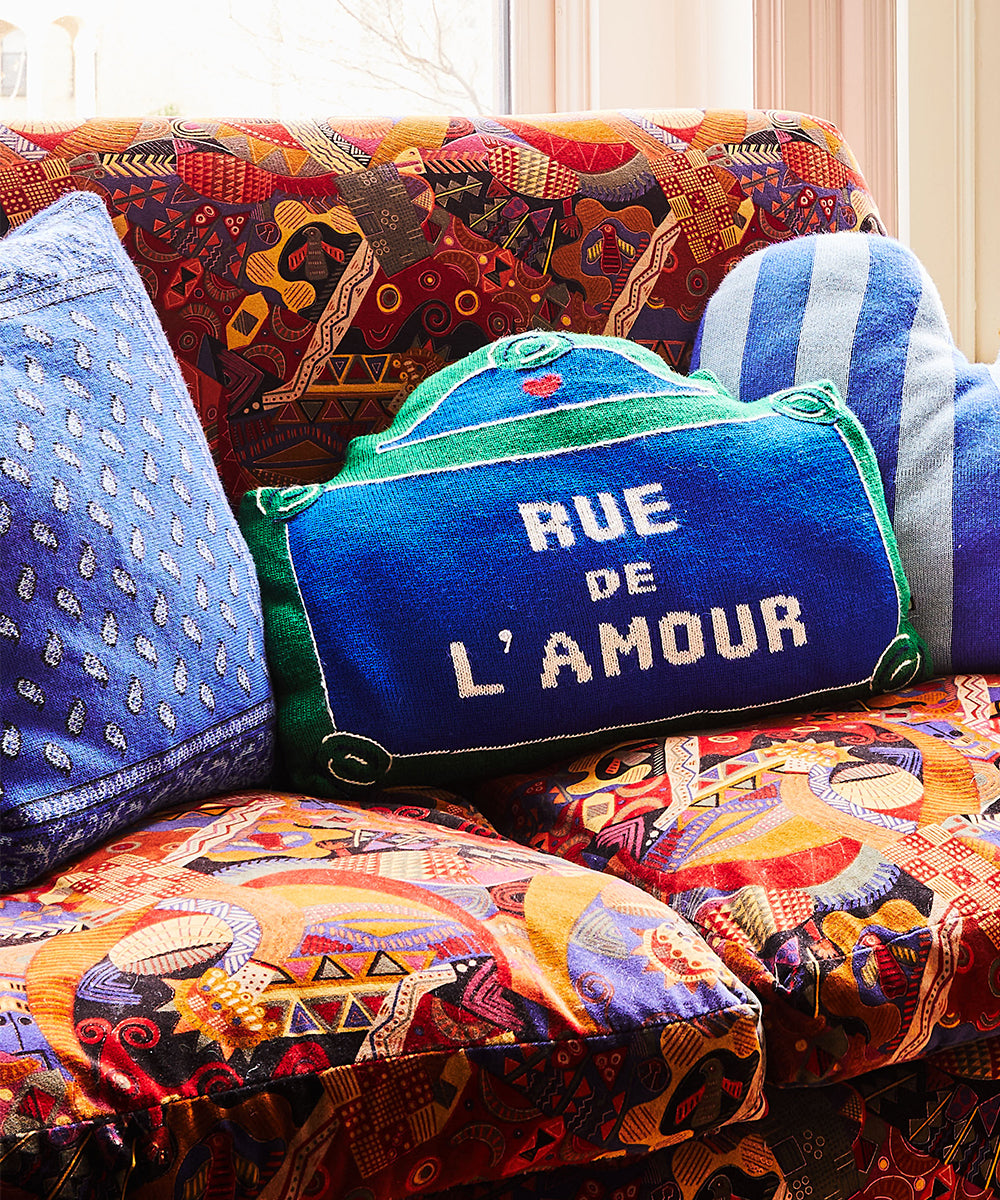 A vibrant Rue de lAmour Pillow adds a splash of color on a patterned sofa.