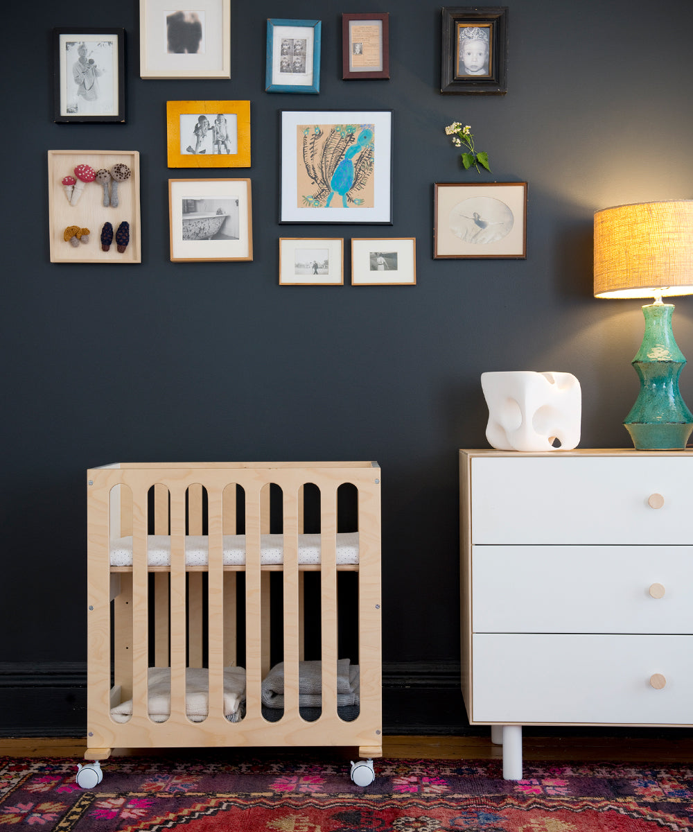 Nursery featuring the Fawn 2-in-1 Crib System, a colorful rug, wall art collage, and a lamp on a dresser.