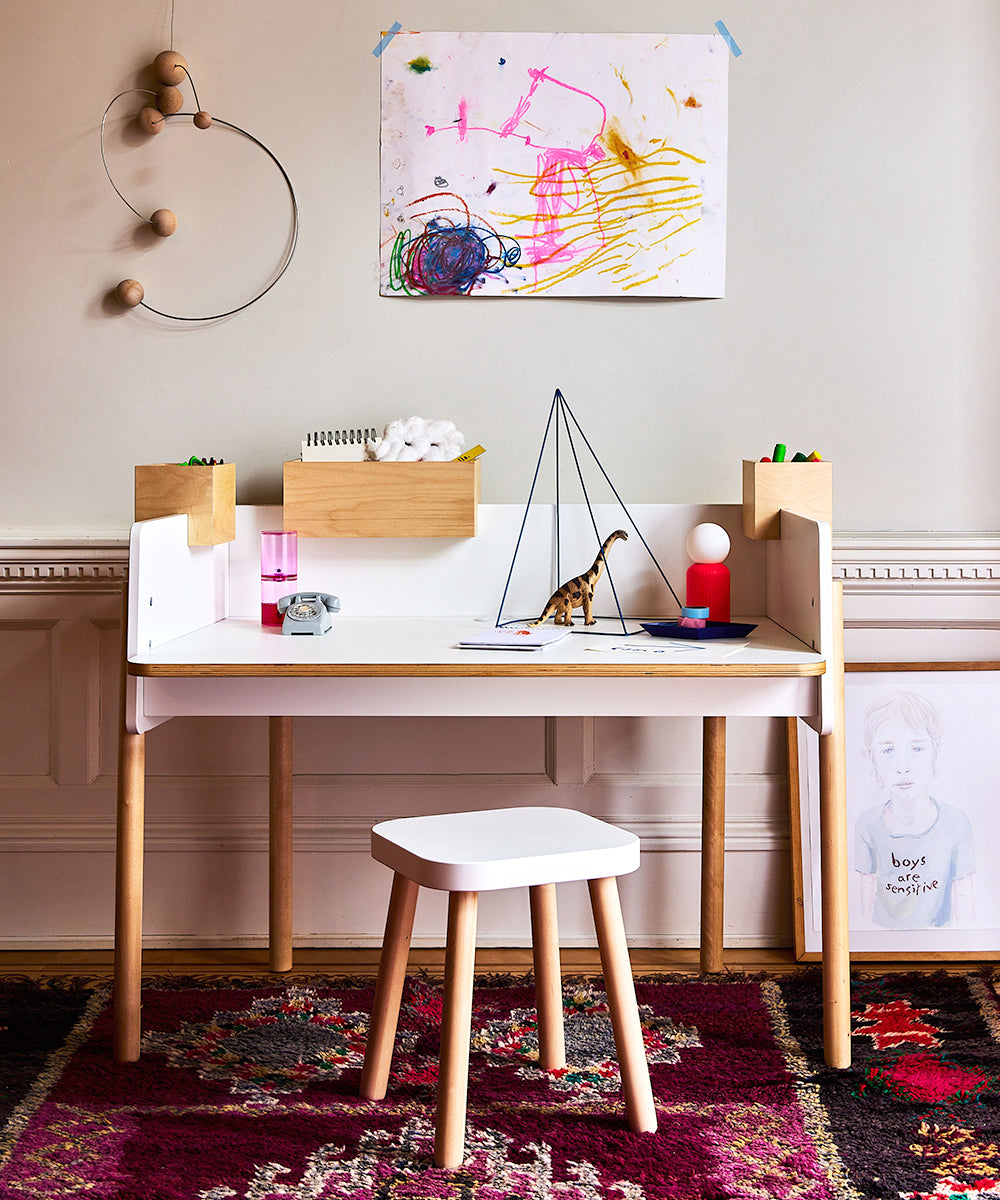 Brooklyn Desk designed for kids features colorful drawings, toys, and includes a stool. It is set against a decorated wall and has a vibrant rug underneath.