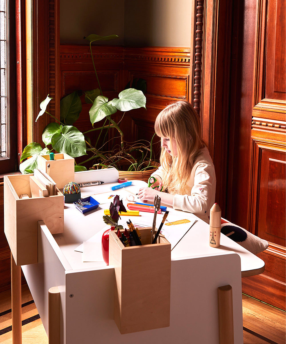 A child is drawing at a Brooklyn Desk with colored pencils, surrounded by wooden walls and a nearby potted plant.