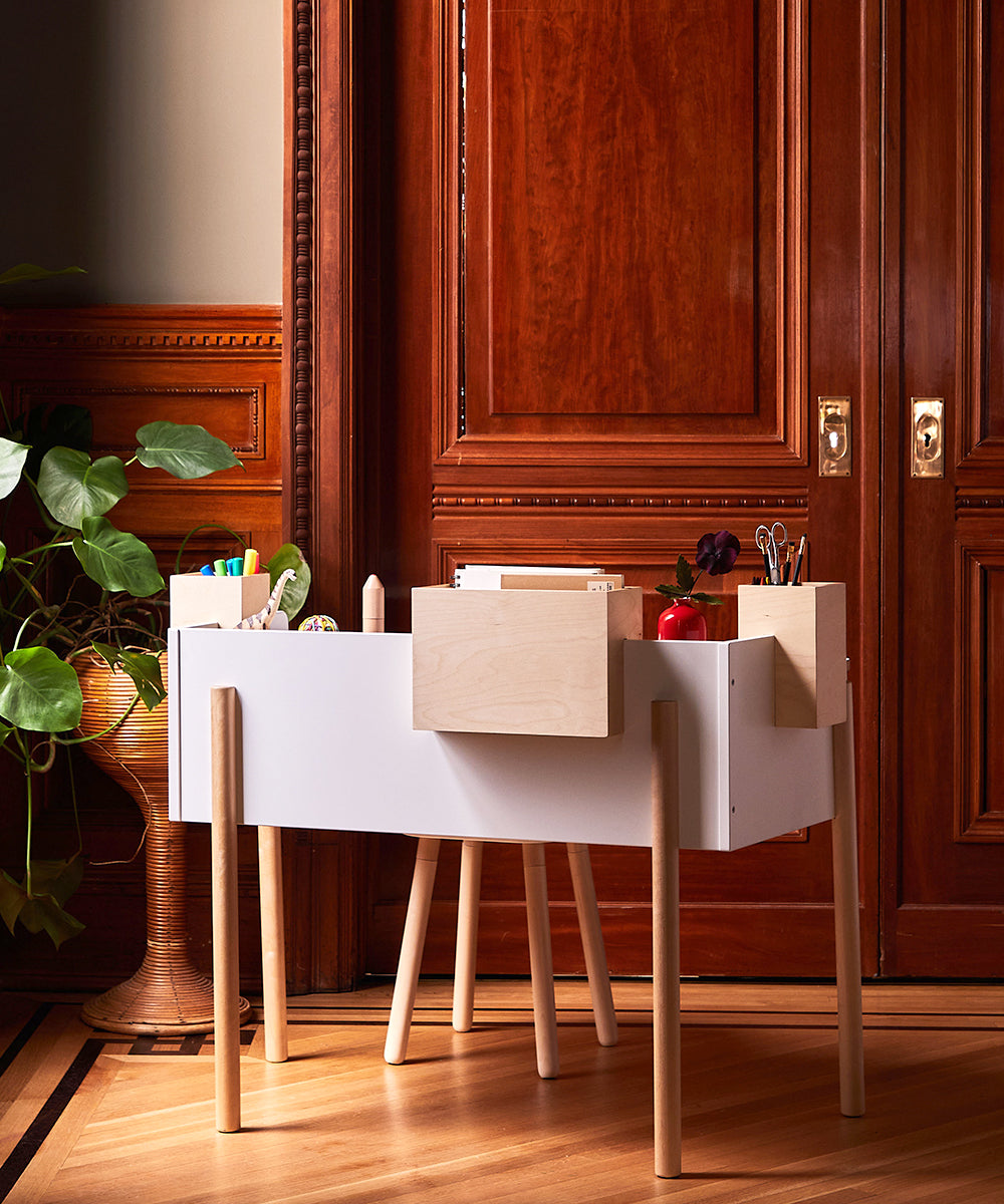 The Brooklyn Desk, a modern white desk with wooden legs, features stationery, a red apple, and decor against a wooden door backdrop.