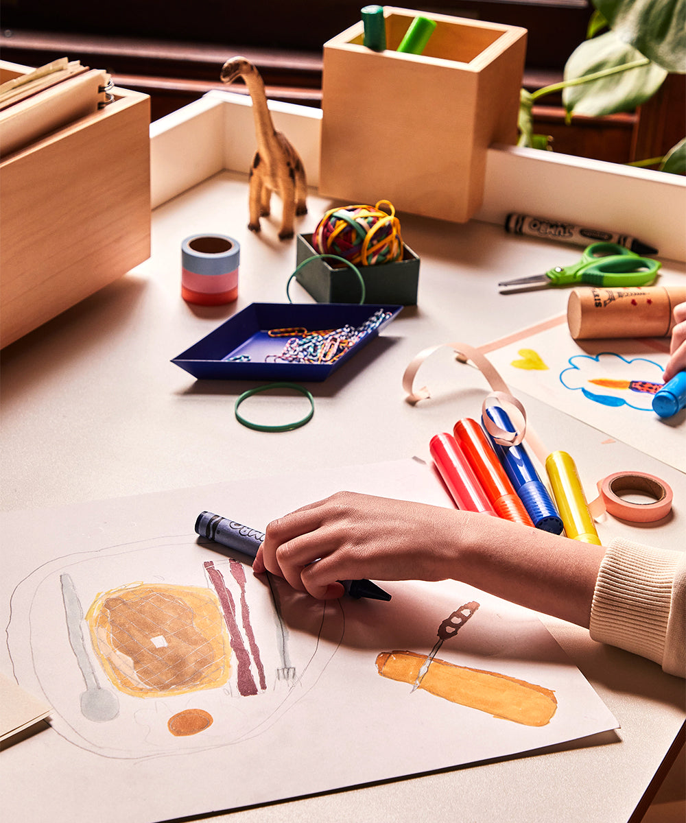 A person draws breakfast items with markers at a cluttered Brooklyn Desk filled with stationery and toys.