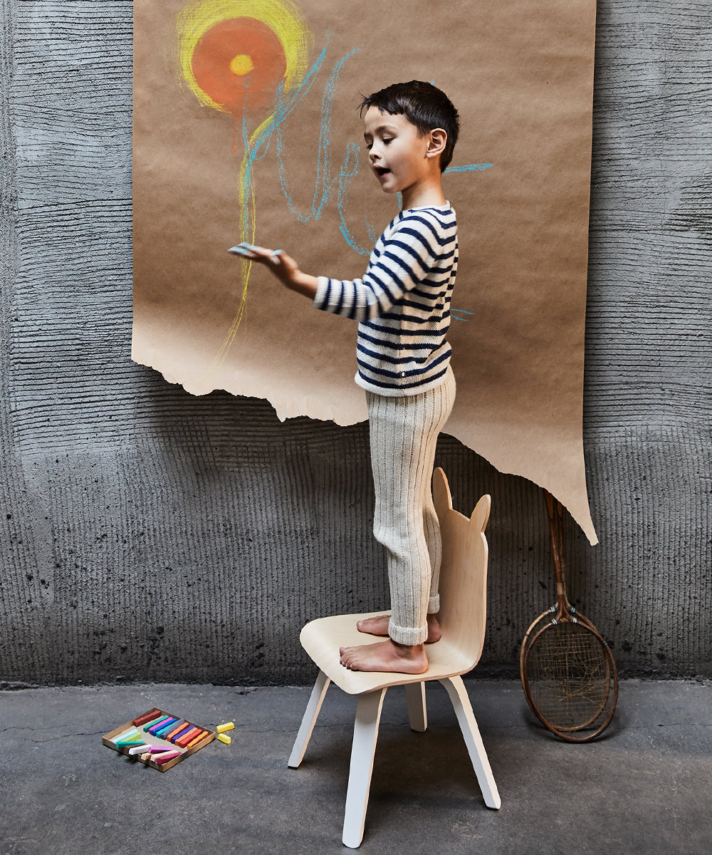 A child in a striped shirt stands on a Bunny Play Chair, drawing with chalk on paper, surrounded by crayons and a tennis racket on the floor.