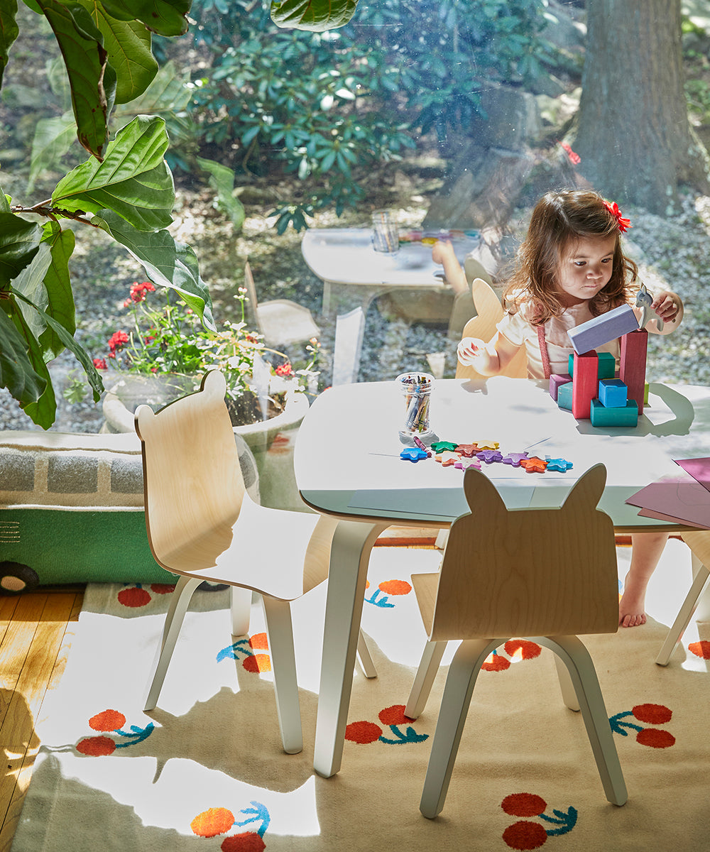 A young girl enjoys playing with colorful blocks at a small table in a sunlit room filled with greenery, seated on the Bunny Play Chair (Set of 2).